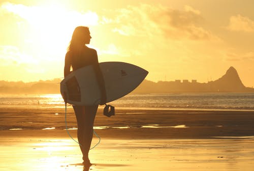 女人抱着冲浪板在日落时站在海岸线上 · 免费素材图片