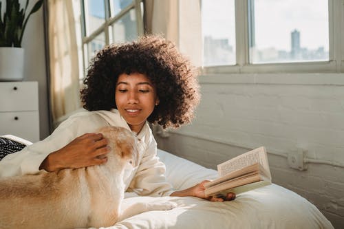 微笑的年轻种族女性爱抚的宠物，当躺在床上和阅读书时 · 免费素材图片