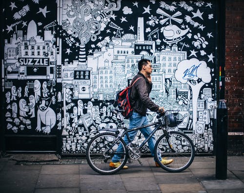 在黑色和白色的艺术墙附近拿着黑色的硬尾自行车的黑夹克的男人 · 免费素材图片