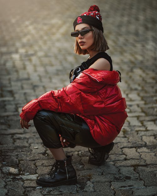 女人穿着红色外套和黑色太阳镜坐在人行道上 · 免费素材图片
