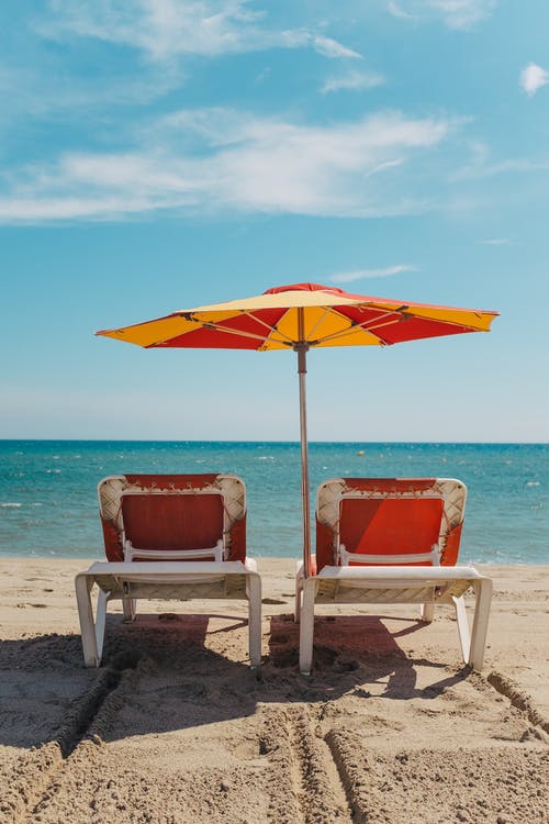 2在海滩上的橙色伞下的红色和白色沙滩椅 · 免费素材图片