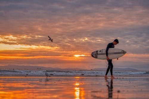 在日落期间携带白色冲浪板在沙滩上行走的人 · 免费素材图片
