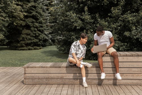 年轻黑人显示笔记本电脑到族裔朋友坐在公园的长椅上 · 免费素材图片