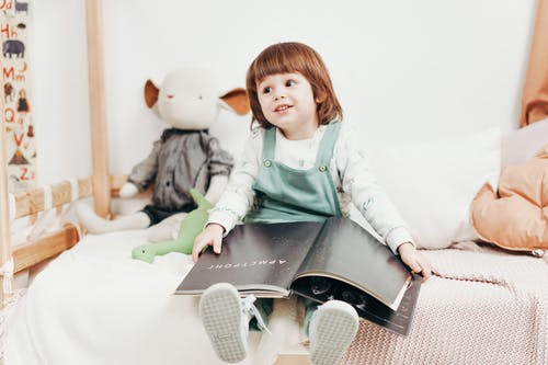 坐在床上拿着本书的白色长袖上衣的孩子 · 免费素材图片