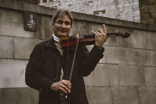 微笑的常设男人，用灰色的石头墙拉小提琴 · 免费素材图片
