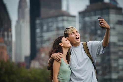 快乐的年轻亚洲夫妇拥抱和微笑，同时在城市采取自拍照 · 免费素材图片