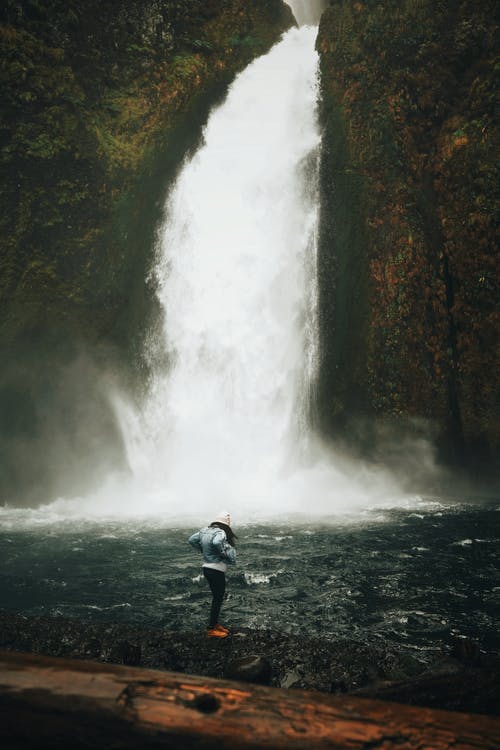 蓝色外套和黑色的裤子，站在瀑布附近的岩石上的人 · 免费素材图片