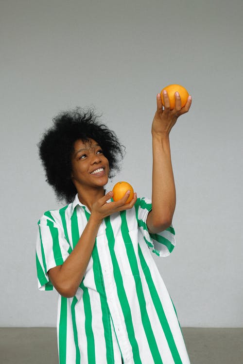白色和绿色的条纹衬衫举起橙色水果的微笑女人的照片 · 免费素材图片