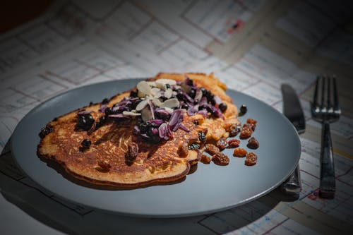 棕色煎饼配棕色紫色白色坚果在银圆刀和叉子旁边的灰色圆板上 · 免费素材图片