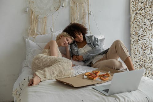 平静的年轻多族裔女友在床上放松的同时吃披萨和看笔记本电脑 · 免费素材图片