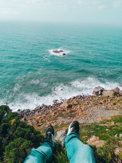 黑色的裤子和黑色的远足鞋，坐在多岩石的海岸上的人 · 免费素材图片