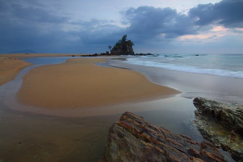 海浪在海边崩溃与岩石形成的看法与树在它之下在灰色云彩和白色天空下 · 免费素材图片