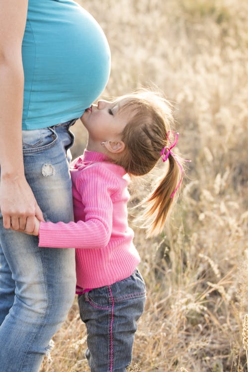 桃红色毛线衣和灰色牛仔裤的女孩亲吻孕妇的肚子在蓝色衬衣和蓝色牛仔布牛仔裤 · 免费素材图片