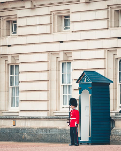 男警卫穿着红色和白色的服装，拿着步枪，而站在白色和蓝色的耳朵棚 · 免费素材图片