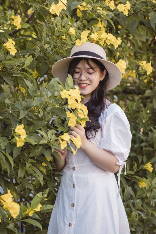 白色的裙子和白色的帽子，站在黄色的花树旁边的女人 · 免费素材图片