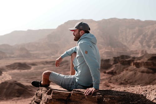 灰色连帽衫和黑帽坐在棕色岩石上的男人 · 免费素材图片