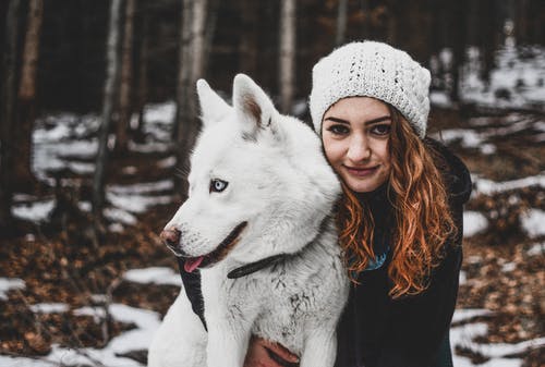 拥抱她的白色西伯利亚雪橇犬时戴着白色针织帽的女人 · 免费素材图片