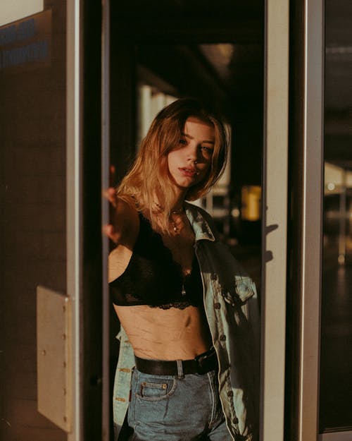 黑色胸罩和蓝色牛仔夹克站在玻璃窗口旁边的女人 · 免费素材图片