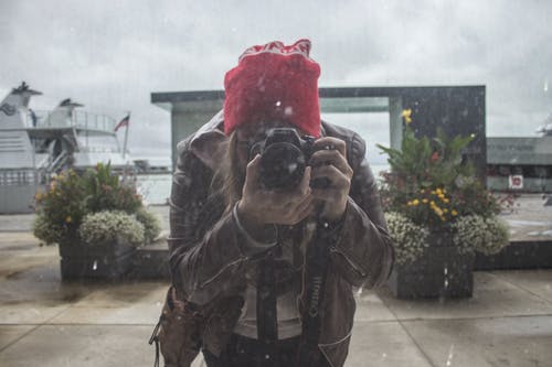 棕色皮夹克和红色针织帽，使用黑色单反相机的女人 · 免费素材图片