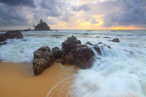 岩石在海滩海岸与海浪在多云的白天天空中崩溃 · 免费素材图片