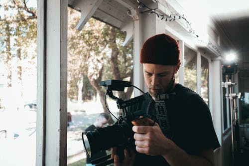 男子携带黑色摄像机站在窗户旁边 · 免费素材图片