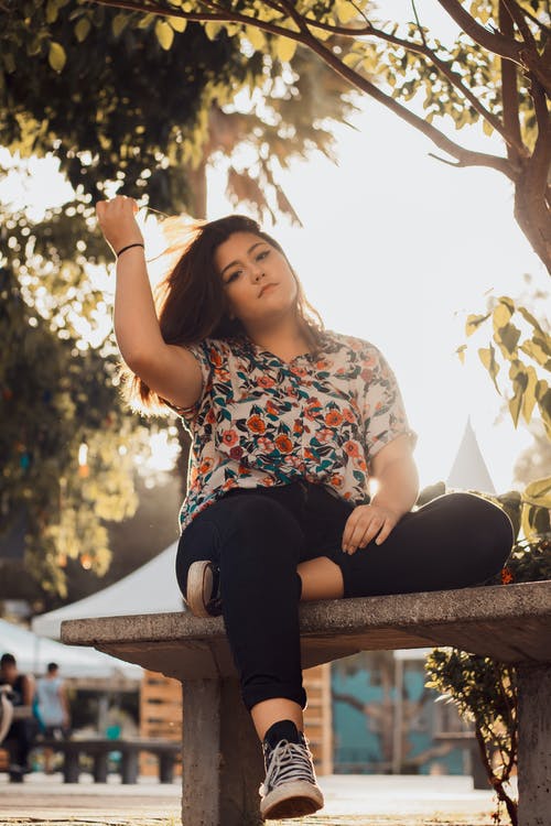 女人穿着花衬衫和黑色牛仔牛仔裤，坐在长椅上 · 免费素材图片
