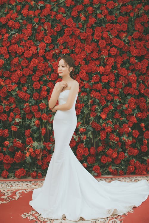 身穿白色甜心领口紧身衣的女人穿着长礼服站在和微笑附近红玫瑰花朵 · 免费素材图片