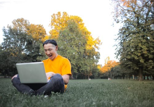 黄船员的脖子t恤和灰色的裤子，坐在绿草地上，使用笔记本电脑的人 · 免费素材图片