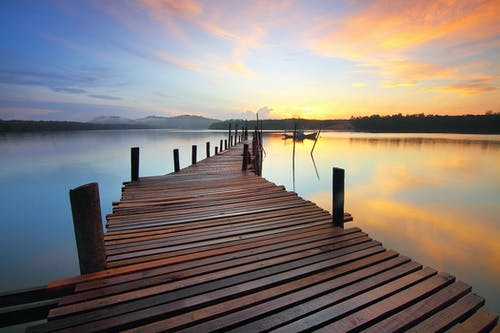 棕色木制码头上平静的水体，日落期间树木的轮廓包围 · 免费素材图片