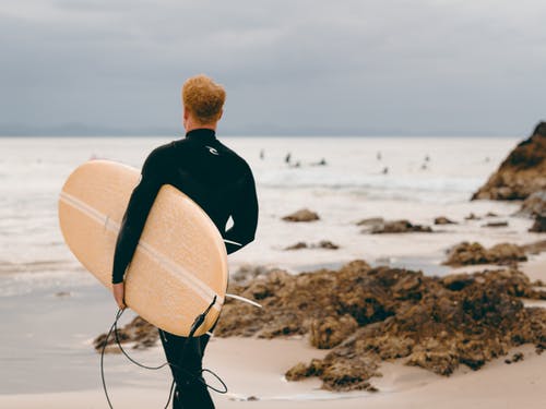 穿米色和白色冲浪板在海滩上的黑夹克的男人 · 免费素材图片