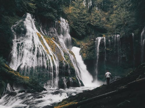 在瀑布前的黑岩石上穿白衬衣的男人 · 免费素材图片