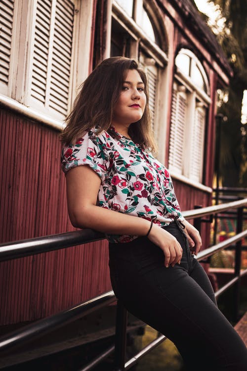 白色蓝色和红色的花衬衫和黑色的裤子，靠着黑色栏杆的女人 · 免费素材图片