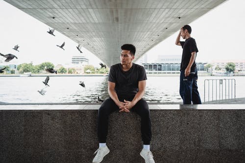 嬉戏的年轻男子坐在花岗岩路堤和朋友站在城市背景后面 · 免费素材图片