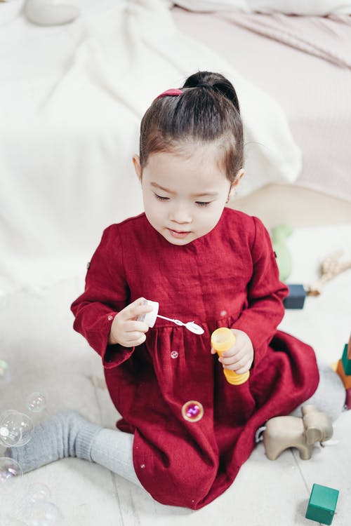 红色长袖穿裙子的女孩坐在白色的地砖，玩吹泡泡机 · 免费素材图片