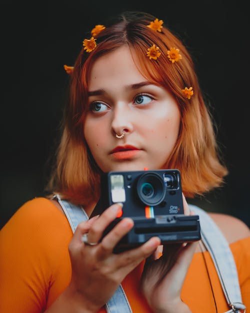 拿着灰色和黑色相机的橙色背心的女人 · 免费素材图片