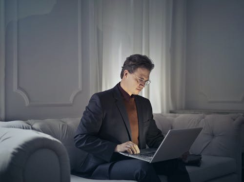 穿着黑色西装，戴着眼镜坐在使用macbook的灰色沙发上的男人 · 免费素材图片