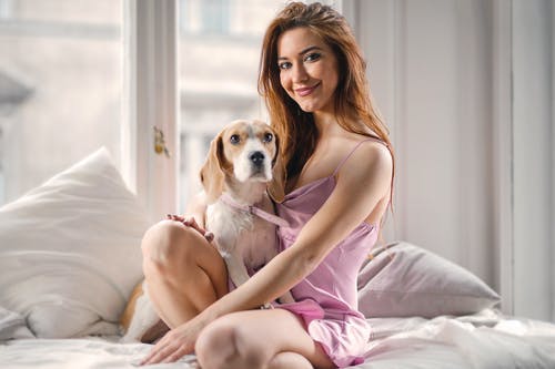 坐在床上与白色和棕色的短涂狗的粉红色裙子的女人 · 免费素材图片