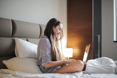 在使用笔记本电脑时坐在床上的黑色和白色条纹顶微笑女人的侧视图照片 · 免费素材图片