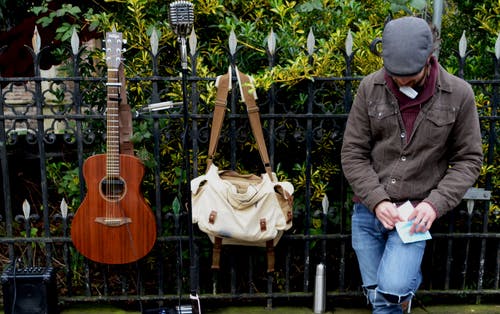 男子倚在白色和棕色的扁带袋和棕色的原声吉他旁边的黑色钢栅栏 · 免费素材图片