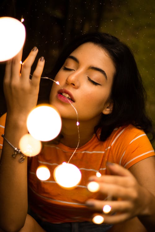 橙色t恤的女人，她的眼睛闭上持有点燃的灯串 · 免费素材图片