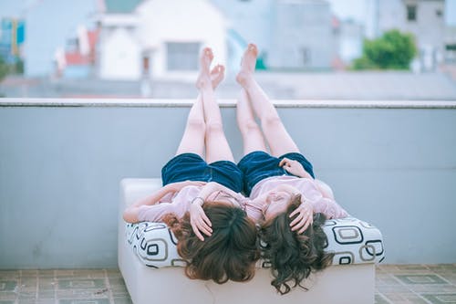 两个女人穿着粉红色的衬衫和蓝色的短裤，躺在沙发上彼此面对，而双脚交叉并放置在混凝土墙上 · 免费素材图片