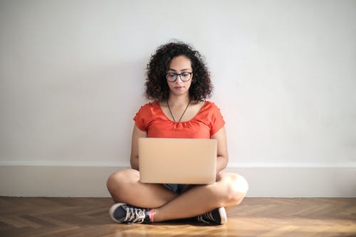 橙色乘员脖子t恤坐在棕色木地板上使用笔记本电脑的女人 · 免费素材图片