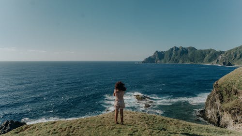 女人穿着白色上衣和短裤站在悬崖附近的水体上的背影照片 · 免费素材图片