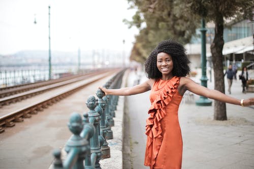 金属围栏摆姿势的橙色无袖连衣裙的微笑妇女的选择性焦点照片 · 免费素材图片