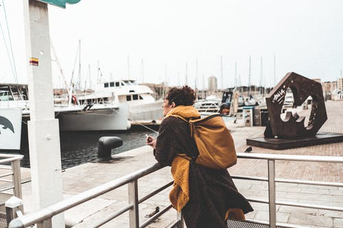 棕色和黑色外套，背着棕色背包，倚在灰色金属栏杆上的女人 · 免费素材图片