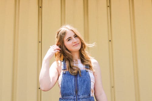 站在棕色墙旁边的蓝色牛仔布的微笑妇女的照片 · 免费素材图片