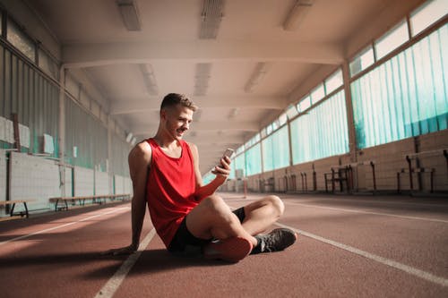 红色背心和黑色短裤的男人坐在跑道上发短信时 · 免费素材图片