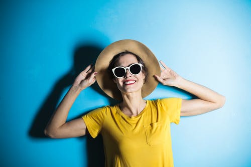 摆在蓝色背景前的黄色t恤，遮阳帽和白色镜框太阳镜的微笑女人的照片 · 免费素材图片