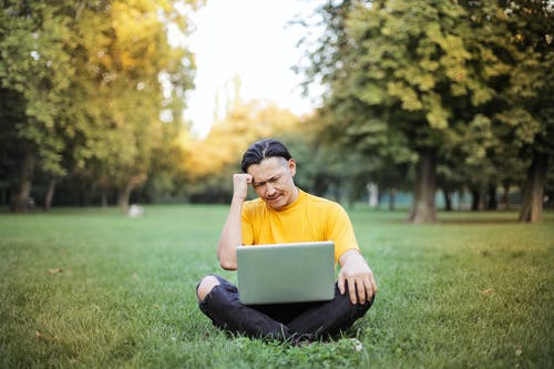 黄色乘员脖子t恤的男人在绿色草地上使用银色macbook · 免费素材图片