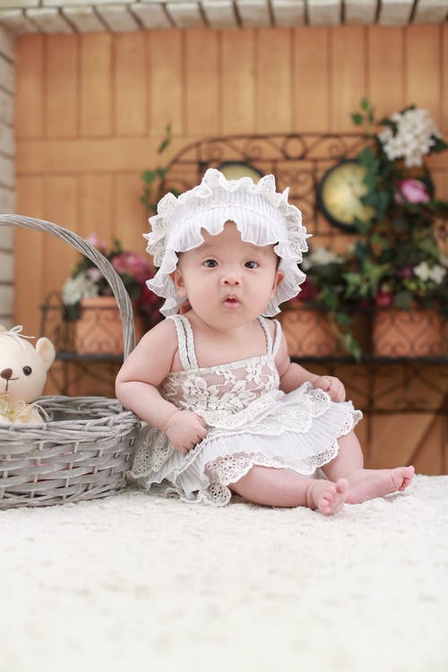 坐在灰色柳条篮子旁边的婴儿头戴白色头带和白色蕾丝花裙子 · 免费素材图片